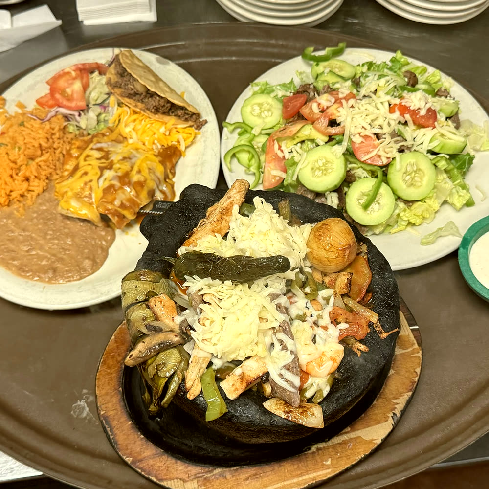 The molcajete: a black stone bowl of grilled meats, shrimp, peppers and melted cheese, beside a cucumber salad and a plate of enchiladas, a crispy taco, rice and beans.