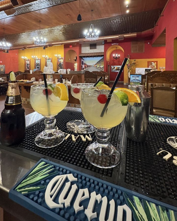 Three lime margaritas with cherries and orange slices on the bar beside a bottle of beer, the red and gold dining room and its chandeliers behind them.